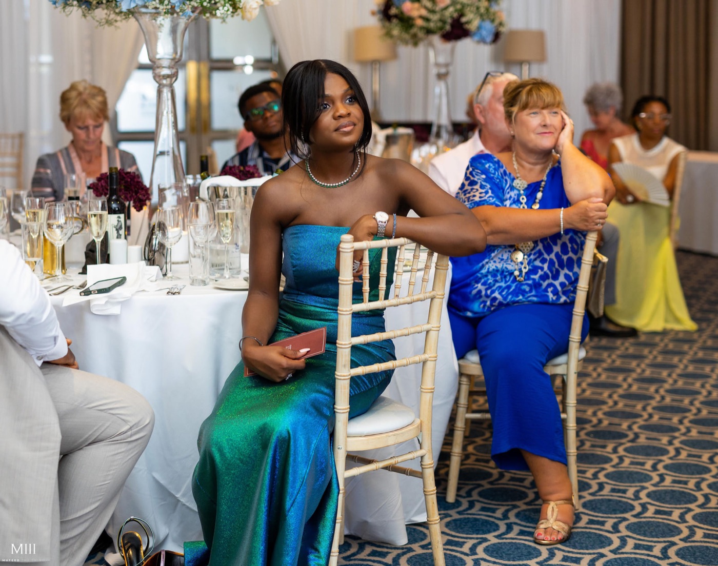 Guests seated at round tables in a hotel ballroom. A young woman in a green gown holds a small card in her lap.